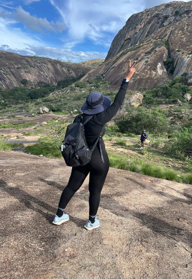 A hiker standing on a rock outcrop with one arm raised, green hills behind her
